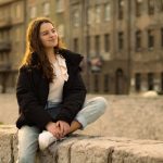 Relaxed young woman with long hair sitting on a stone wall in a cityscape.